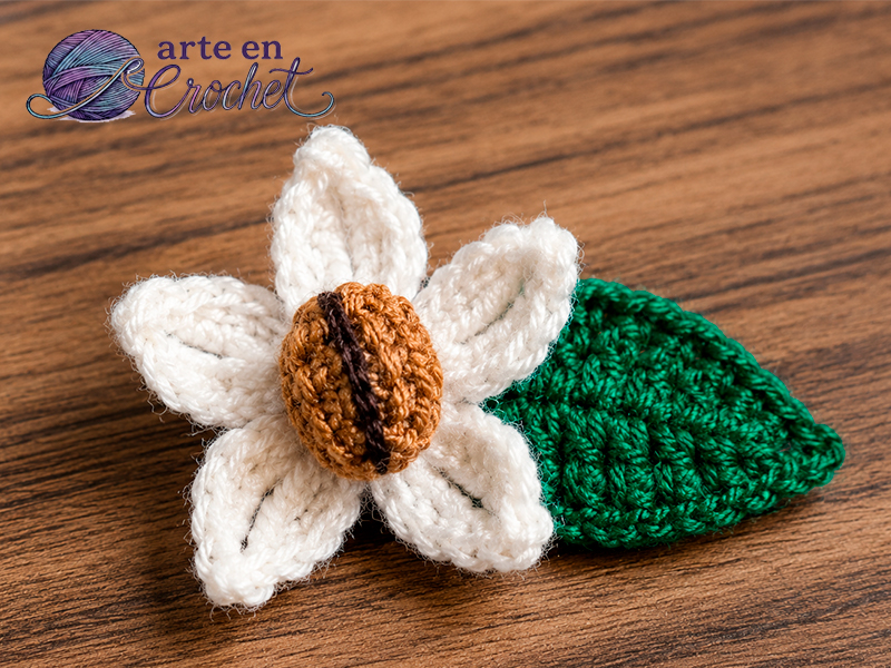 Prendedor de café a crochet con flor blanca, hoja tejida y grano de café tostado, ideal como accesorio artesanal.